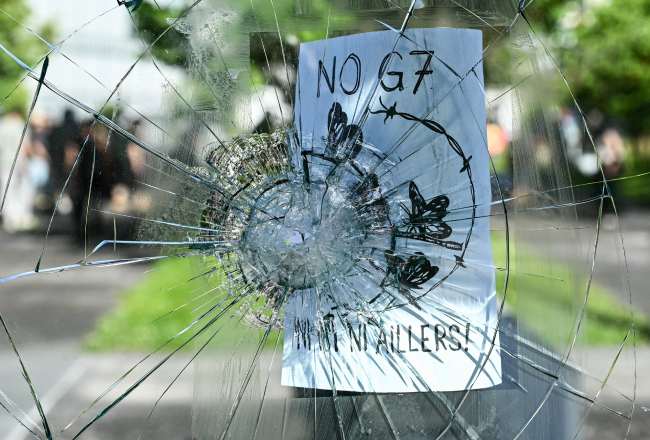 This photograph shows a broken glass with a flyer reading "No G7, not here, not elsewhere" during a rally of a "No-G7" coalition of over 60 associations, unions and left-wing groups "against fascism and imperialism”, a day ahead of the summit's start, in Geneva on June 14, 2026.(Photo by FABRICE COFFRINI / AFP)
